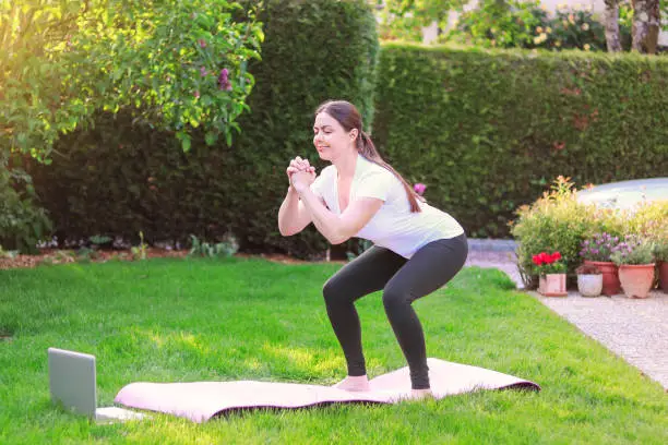 Photo of Healthy lifestyle. Exercising at home. Doing fitness online. Beautiful young woman doing sport in garden outdoors following guide of online tutorial or trainer on laptop.
