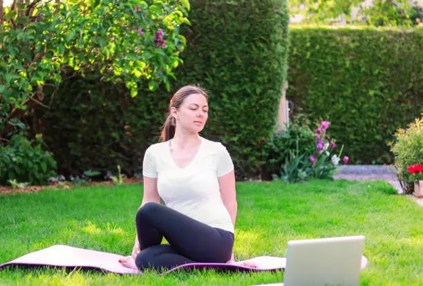 Photo of Beautiful young woman practicing yoga in garden outdoors following guide of online tutorial or trainer on laptop. Healthy lifestyle. Exercising at home. stretching muscles