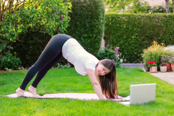 Photo of Beautiful young woman practicing yoga in garden outdoors following guide of online tutorial or trainer on laptop. Healthy lifestyle. Exercising at home. stretching muscles