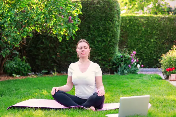 Photo of Beautiful young woman practicing yoga in garden outdoors following guide of online tutorial or trainer on laptop. Healthy lifestyle. Exercising at home. Meditation online.