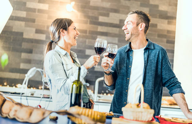 young couple in love drinking red wine at house kitchen - happy millenial people at home enjoying aperitif time cheering together at jubilee anniversary - genuine youth concept on bright indoor filter - wine dinner party drinking toast imagens e fotografias de stock