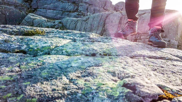A close up on a male hiking boots. The man is walking front. Soft sunrise beams are lighting the way. Rocks overgrown with moss. Barren and empty landscape.