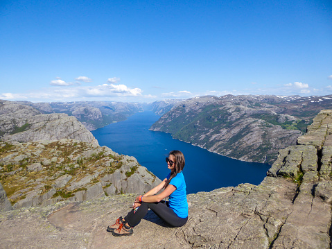 A girl wearing blue T-shirt sitting at the edge of a steep cliff of Preikestolen, with a view on Lysefjorden. Fjord goes far inland. Girl enjoys the view, feeling free and happy. Great accomplishment