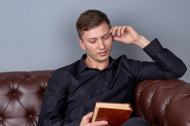 istock Young man is sitting on a leather sofa and reading a book 1150765672