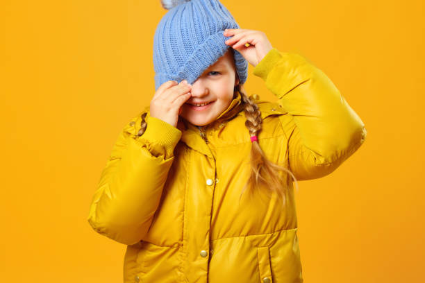 nahaufnahme eines fröhlichen kleinen mädchens in jacke über gelbem hintergrund. das kind schaut unter den hut. - jacket child clothing fashion stock-fotos und bilder