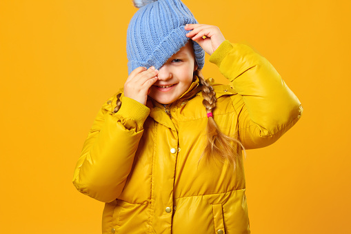 Closeup portrait of a cheerful little girl in jacket over yellow background. The child looks out from under the hat.
