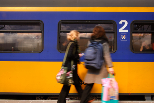 A dslr photo of two woman running to fetch the train. The typical dutch railways yellow and blue train is waiting at the platform. The woman are carrying shopping bags and are motion blurred.