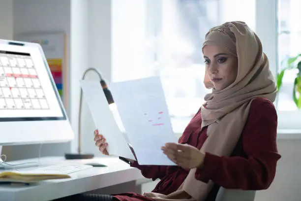 Sitting near computer. Dark-eyed teacher wearing hijab sitting near computer checking her week schedule