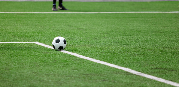 Soccer ball on the white line on the grass, which marks the area of the soccer field. In the blurred background, the feet of a soccer player.