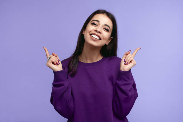 pretty adult girl with broad smile, wears purple sweatshirt, pointing fingers up at copyspace, isolated over violet wall - camisa de moleton imagens e fotografias de stock