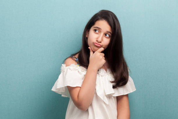 portrait of thoughtful beautiful brunette young girl with black long straight hair in white dress standing, touching her chin, looking away and thinking. - pensive question mark teenager adversity imagens e fotografias de stock