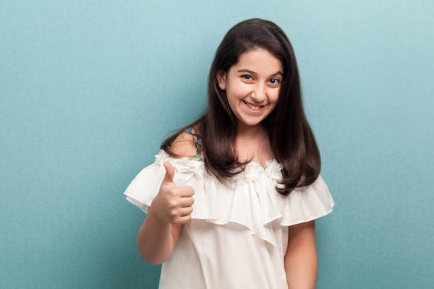 porträt der glücklichen zufriedenen schönen brünette junge mädchen mit schwarzen langen geraden haaren in weißem kleid stehen, daumen nach oben und blick auf die kamera. - thumbs up human thumb human finger brown hair stock-fotos und bilder