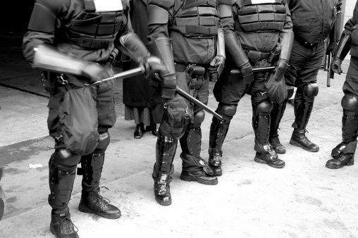 Police bodies in riot gear. Action shot (leftmost officer is smacking his club into his hand in warning). Black and white.