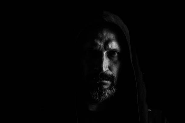 istock Brutal man with a gray beard in the hood with sharp shadows on a black background. Copy space. 1150861072