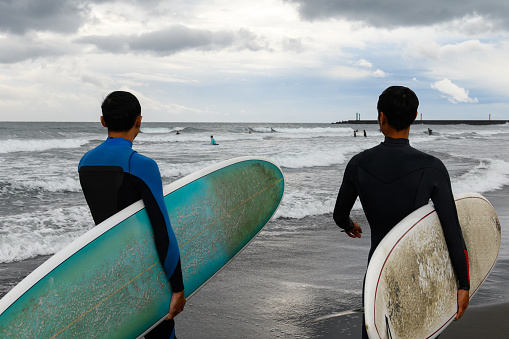 Rear view of determined surfers standing on shore. Male friends are carrying surfboards while looking at sea view at beach. Men are wearing wetsuits while anticipating against cloudy sky.
