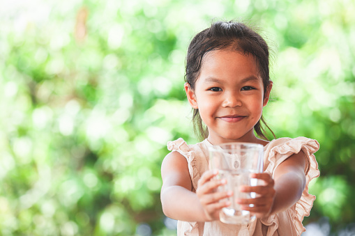 Cute asian child girl like to drink water and holding glass of fresh water in green nature background