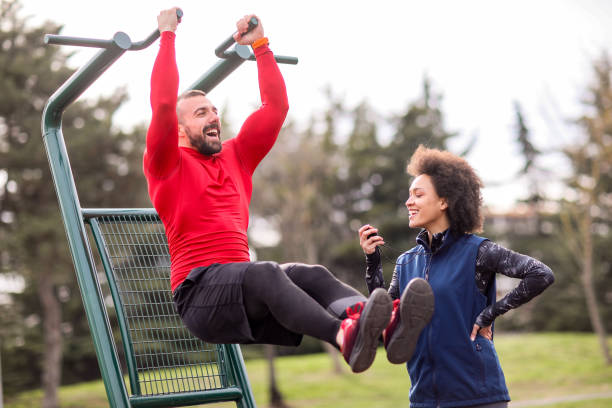 istock Strong male athlete doing a lower abs workout on a pull-up bar in a city park with his female coach standing next to him and measuring time 1150906332