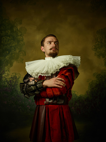 Young man as a medieval knight on dark studio background. Portrait in low key of male model in retro costume. Looks serious. Human emotions, comparison of eras and facial expressions concept.