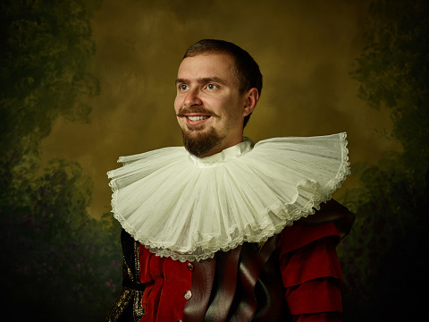 Young man as a medieval knight on dark studio background. Portrait in low key of male model in retro costume. Standing and smiling. Human emotions, comparison of eras and facial expressions concept.