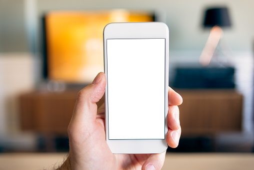 Home Multimedia - User point of view (POV) holding up a smart phone, interacting with TV in the background. The mobile phone has a blank white screen to drop-in your graphic.