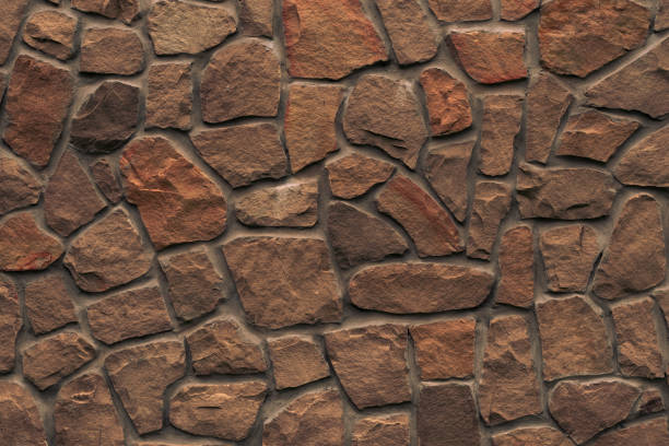 Orange stone wall. Bright brown rock texture. Red faceted stone wall background for design. Modern architecture, granite structure. Red brick wall grunge background. Cement construction material texture. Stonework, lithic, crushed stone