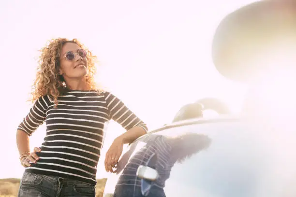 Photo of Beautiful curly blonde cute middle age woman in outdoor happy activity looking at the sun during a bright golden sunset - caucasian female with convertible car for vacation concept