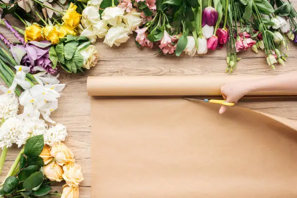 Photo of Partial view of florist cutting craft paper with scissors near flowers on wooden table