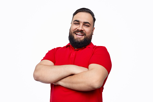 Plus size bearded ethic man in red T-shirt keeping arms crossed and cheerfully smiling for camera while standing against white background