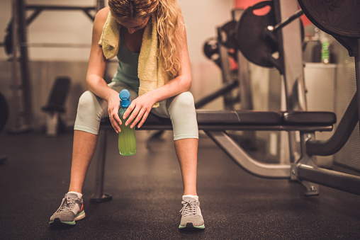 Young blondie woman drinking water and sitting on bench in gym.