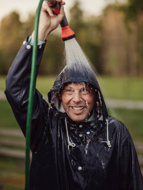 mann schafft regen mit gartenschlauch im regenmantel - drenched raincoat rain clothing stock-fotos und bilder
