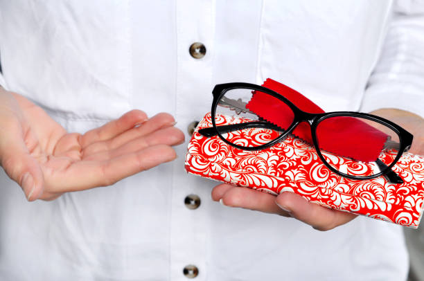 Ophthalmologist holding eyeglasses for a try out. Optometrist hands holding new black plastic eyeglasses red microfiber on white-red case, at optician shop.Close-up view focused on glasses.