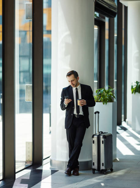 istock Middle adult businessman in suit using phone and drinking takeaway coffee while leaning on column near window in airport waiting for flight with his trolley bag nearby 1150997305