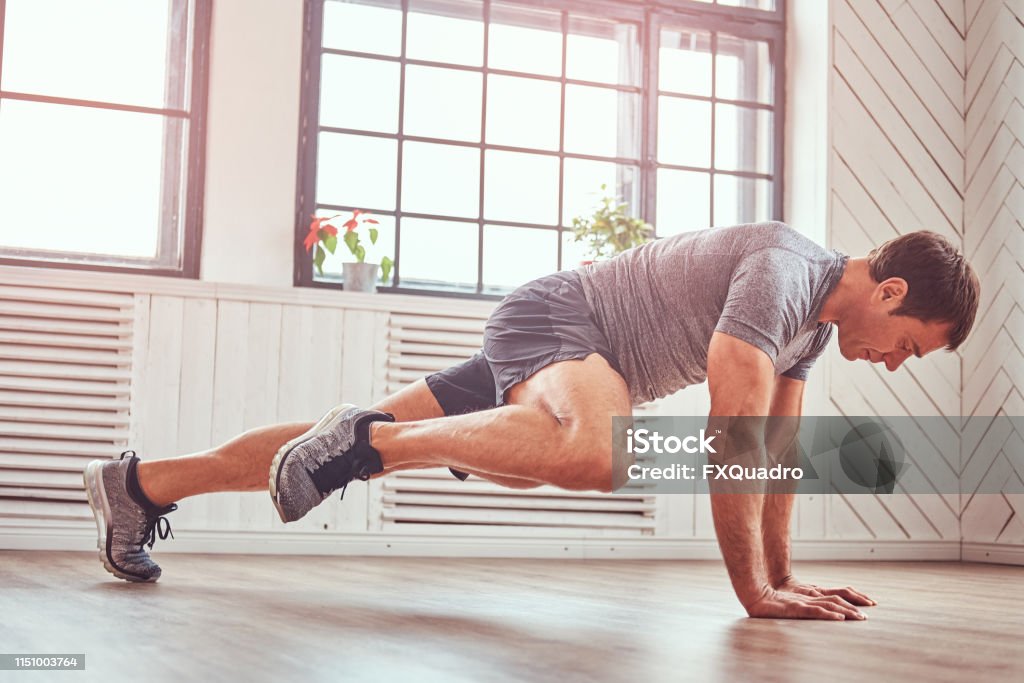 Hombre musculoso guapo en una camiseta y shorts haciendo ejercicios funcionales en el piso en casa. - Foto de stock de Ejercicio físico libre de derechos