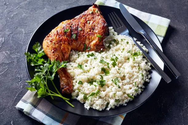 Crispy grilled Chicken Leg Quarter served with Cauliflower rice or couscous on a black plate on a concrete table, horizontal view from above