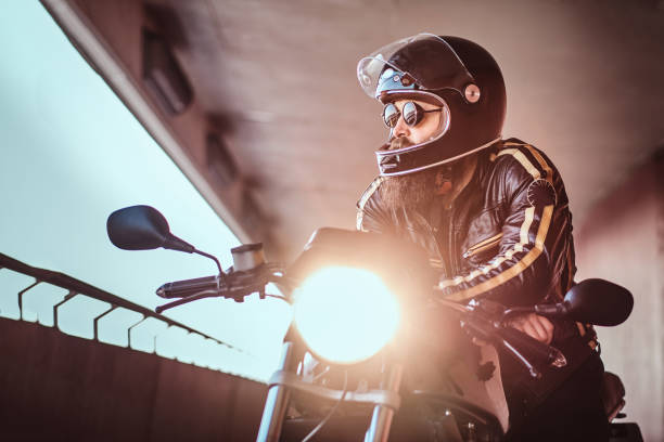 ritratto ravvicinato di un brutale motociclista barbuto con casco e occhiali da sole vestito con una giacca di pelle nera seduta su una moto retrò con un faro incluso. - under attack foto e immagini stock