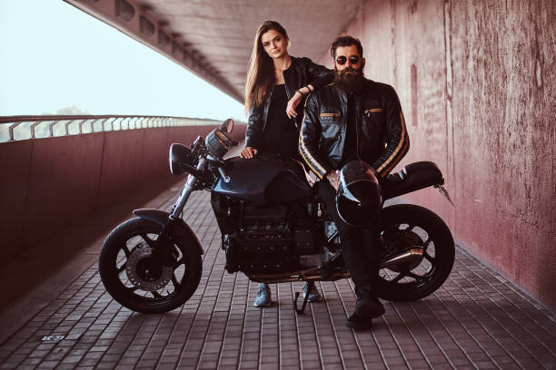 Portrait of a brutal fashionable biker dressed in a black leather jacket with sunglasses sitting on his custom-made retro motorcycle and a young sensual brunette girl who leaning on his shoulder. stock photo