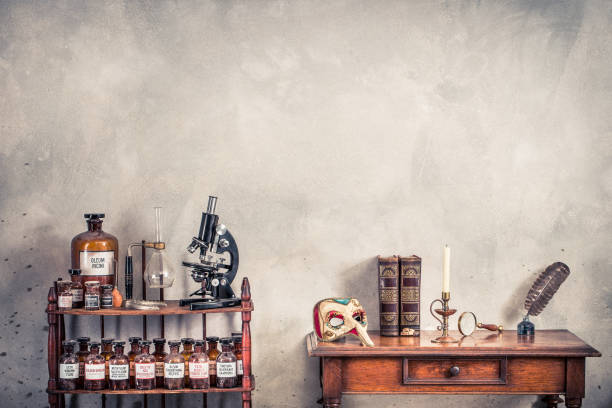 microscopio antiguo, viejas botellas de laboratorio en estanterías de madera, máscara de carnaval, pluma de tinta de quill, inkwell, libros, lupa, vela en candelabro vintage en mesa de roble. foto filtrada de estilo retro - vanitas fotografías e imágenes de stock