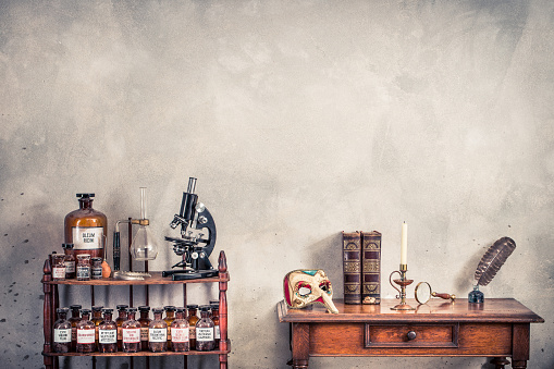 Antique microscope, old laboratory bottles on wooden shelving, carnival mask, quill ink pen, inkwell, books, magnifying glass, candle in vintage candlestick on oak table. Retro style filtered photo