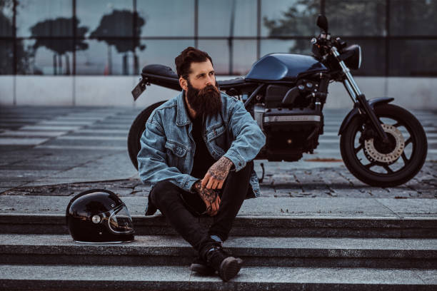 Portrait of a brutal bearded male dressed in a jeans jacket sitting on steps near his custom-made retro motorcycle against skyscraper. Portrait of a brutal bearded male dressed in a jeans jacket sitting on steps near his custom-made retro motorcycle against a skyscraper. denim jacket stock pictures, royalty-free photos & images