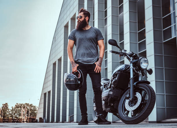 Brutal bearded male in sunglasses dressed in a gray t-shirt and black pants standing near his custom-made retro motorcycle against skyscraper. Brutal bearded male in sunglasses dressed in a gray t-shirt and black pants standing near his custom-made retro motorcycle against a skyscraper. biker stock pictures, royalty-free photos & images