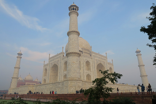 Agra, Uttar Pradesh, India - February 13, 2019:  Stock photo showing Agra’s world-famous Mughal Taj Mahal, an imposing tomb mausoleum which dates back to the 17th century. The elaborate inlaid white marble architecture facade is flanked by four lighthouse-shaped towering minarets, being crowned by a series of large onion domes. Located in Agra, Uttar Pradesh, India, this Taj Mahal photo was taken early in the morning, at sunrise, with crowds of tourists taking selfies and photographs on their mobile phones.