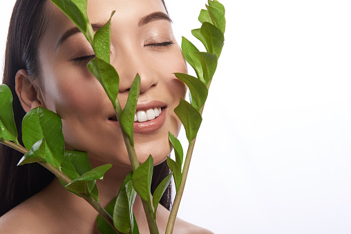 Relaxing with nature. Glad young Asian woman being isolated on the white background and smiling while enjoying leaves near her face