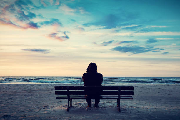 istock Man in hood sitting on a lonely bench on the beach 1151039738