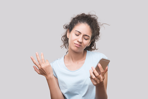 Portrait of sad young woman holding a smart phone over gray background. Horizontal, studio shot.