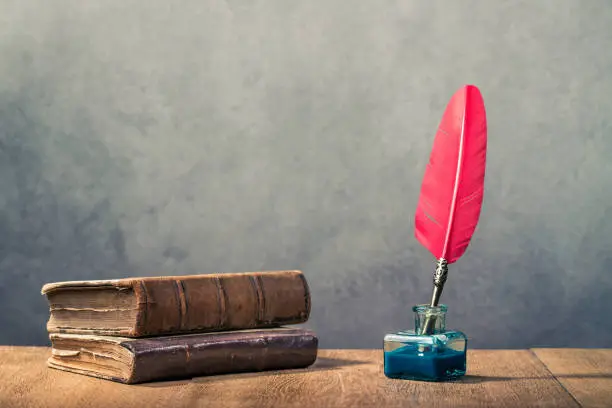 Vintage red quill pen with inkwell and old books on wooden table front concrete wall background. Retro instagram style filtered photo