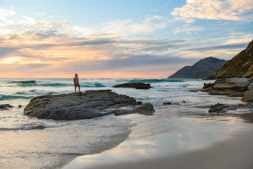 A caucasian, American man in his 30s stands on top of a rock along the scenic landscape of Noordhoek Beach at dusk in Cape Town South Africa.
