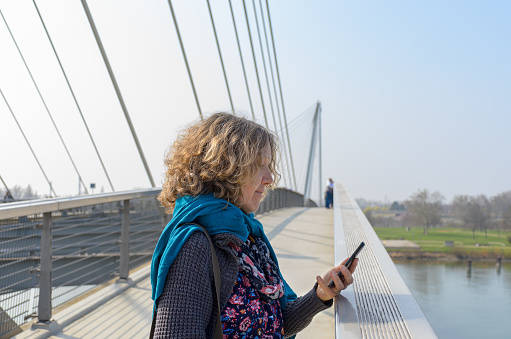 Woman with curly hair standing on modern pedestrian bridge with cell phone in her hand, looking at the screen. Side half-length portrait