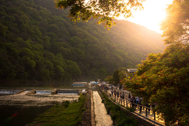 The Hozugawa River winds its way from the lush green mountains of Kameoka to Arashiyama in Kyoto, Japan. Shot from Togetsukyo Bridge in spring at sunset, some kudari tour boats can be seen docked in the distance. Tourists enjoy taking a stroll along the river banks.