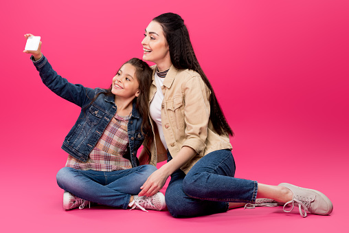happy mother and daughter sitting together and taking selfie with smartphone on pink
