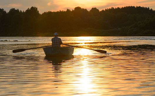 The Caucasian old fisherman is in a boat at sunset. The elderly man is fishing and rowing with oars in a river.